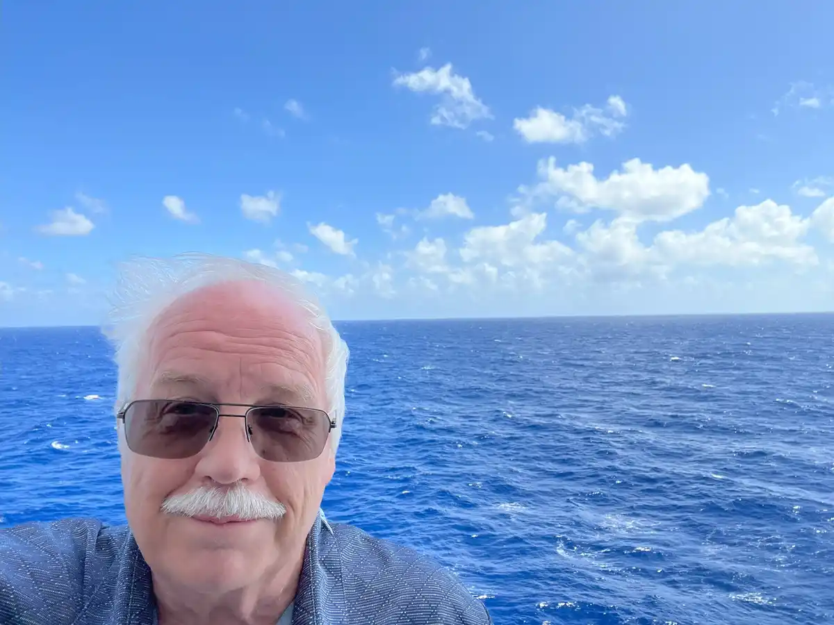 A selfie of an older man with white hair and a mustache wearing sunglasses, smiling at the camera against a backdrop of a…