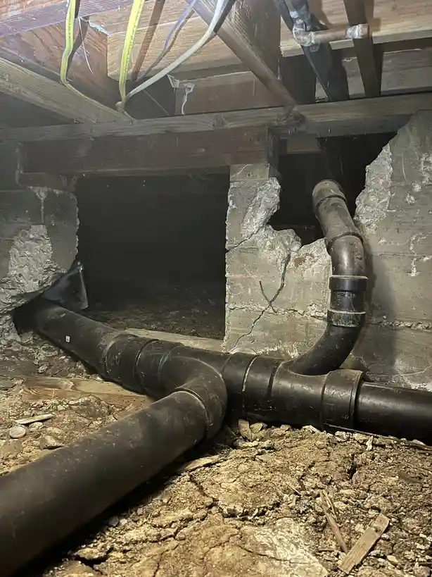 A dimly lit crawlspace shows large black pipes running across dirt and debris, with cracked concrete walls and wooden beams…