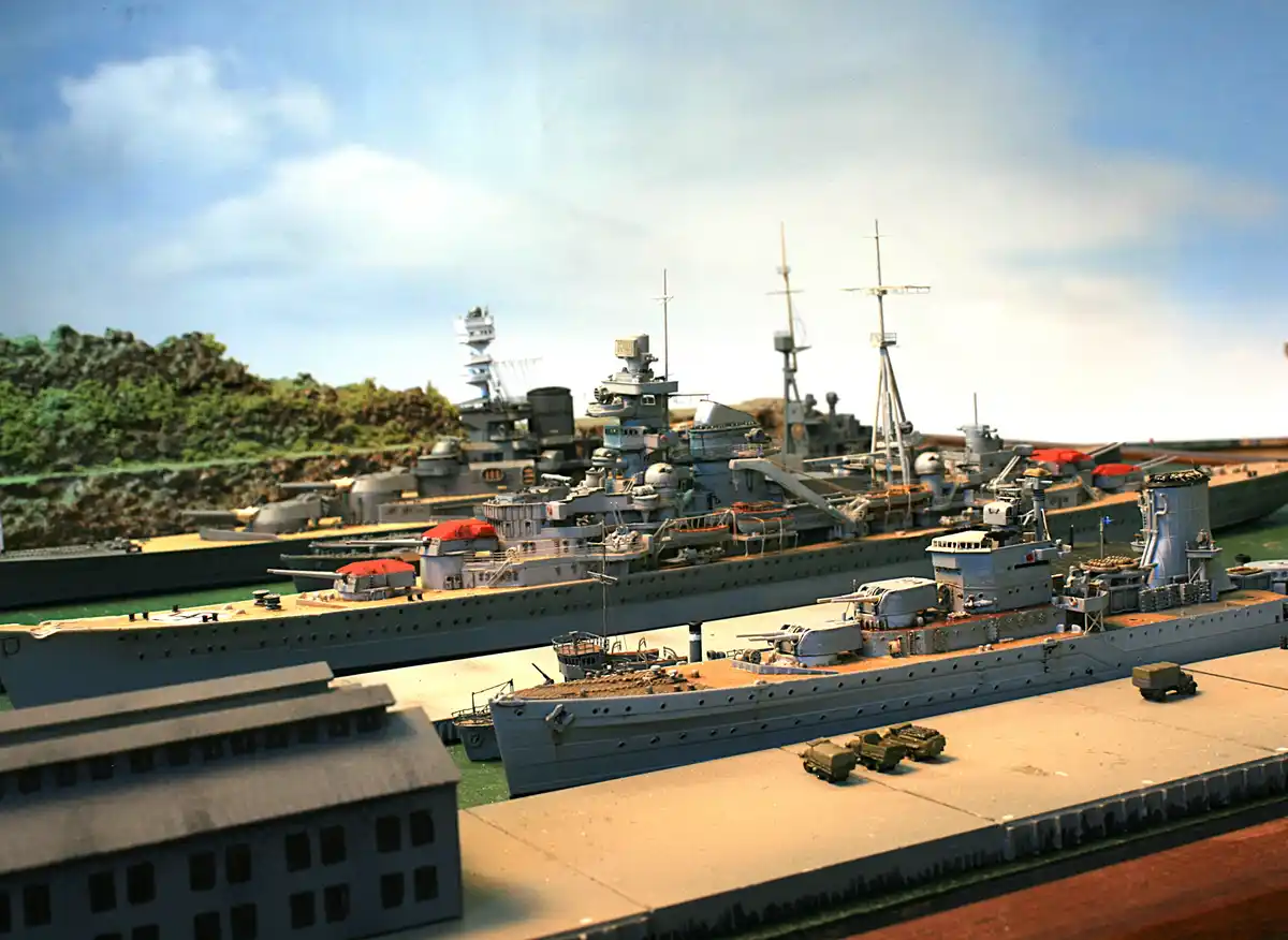 A detailed diorama of several grey warship models docked in a harbor, with miniature buildings and trucks nearby. The…
