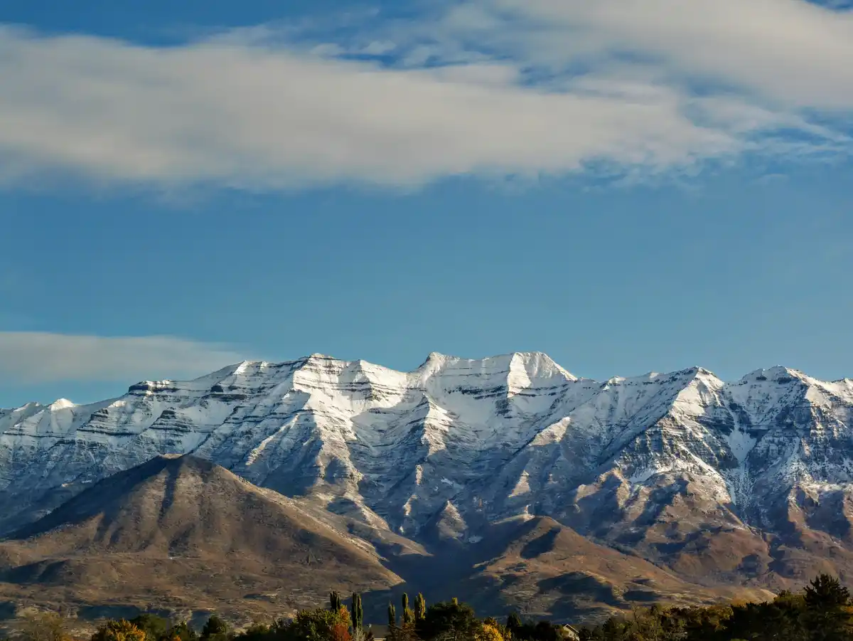 A scenic photo of snow-capped mountains under a blue sky with wispy clouds, with a foreground of autumn-colored trees and…