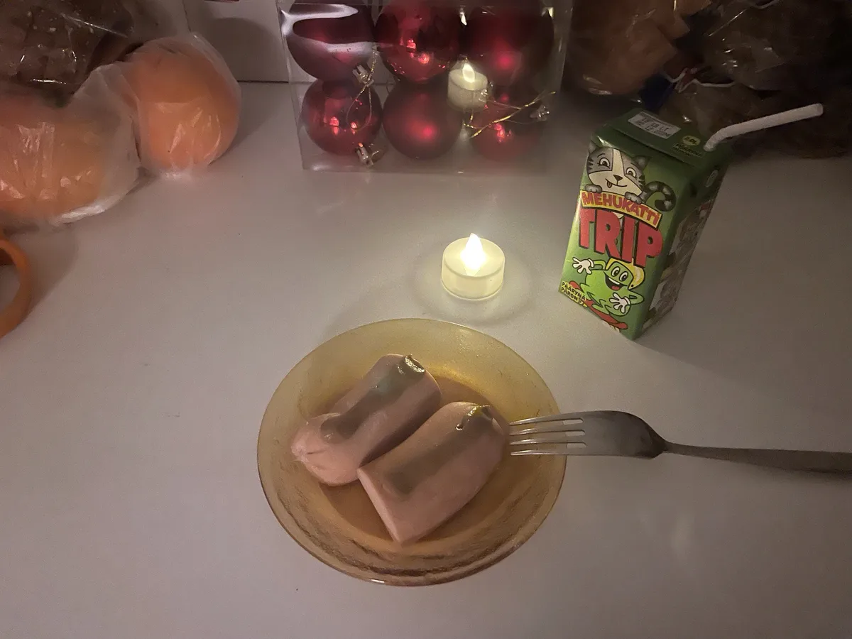 A dimly lit table scene features two pink, sausage-like food items on a yellow plate with a fork, next to a lit tea light…