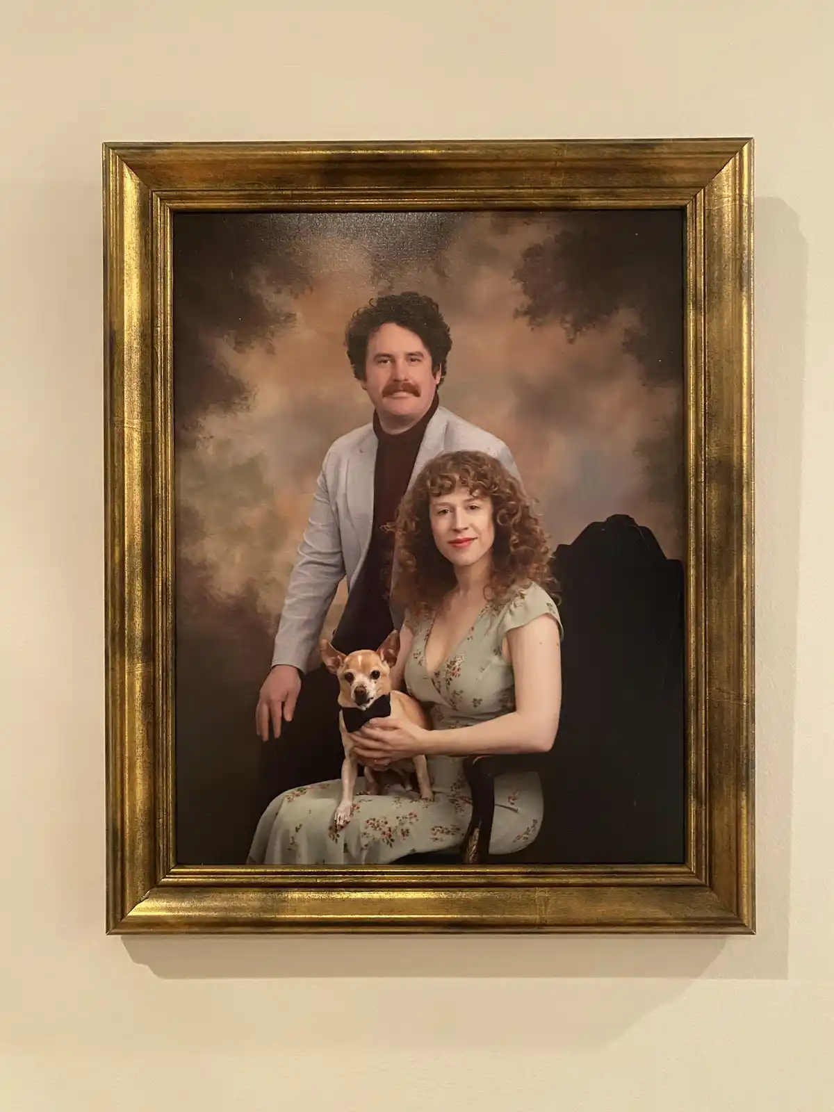 A framed, professionally styled portrait depicts a man with a mustache, a woman with curly hair, and a small chihuahua…