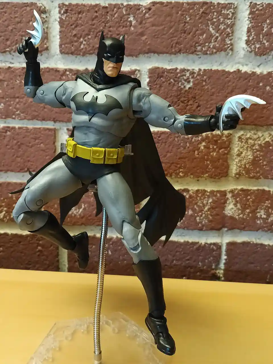 A Batman action figure in a grey and black suit with a yellow utility belt stands in a dynamic pose against a brick wall…