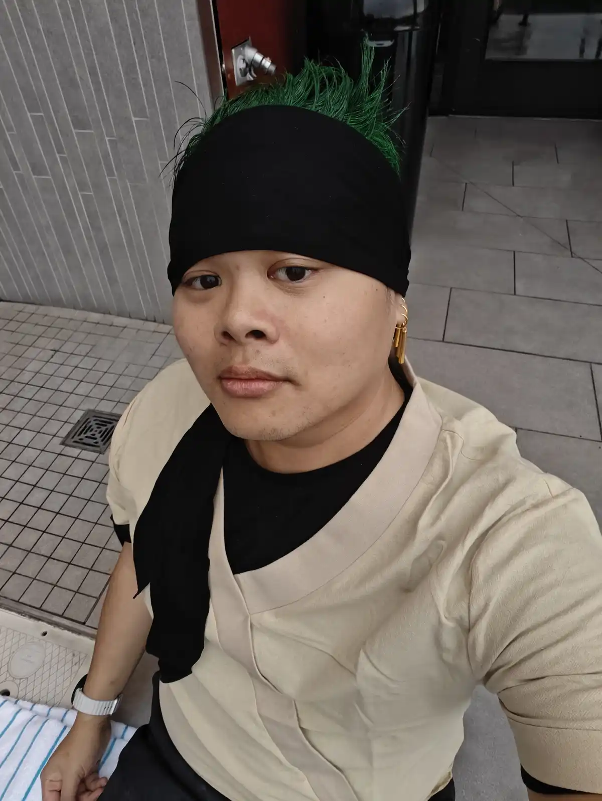 A person with green mohawk hair and a black headband sits outdoors, wearing a beige top over a black shirt, gold earrings…