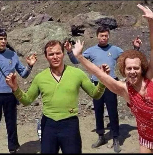 Four men in Star Trek uniforms stand outdoors on rocky terrain, with one man in a green shirt in the center looking annoyed…