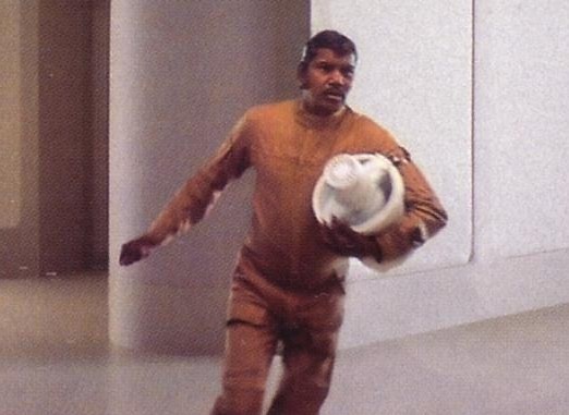 A man in a brown jumpsuit is running while holding a white, round object resembling a helmet or dome. He has a serious…