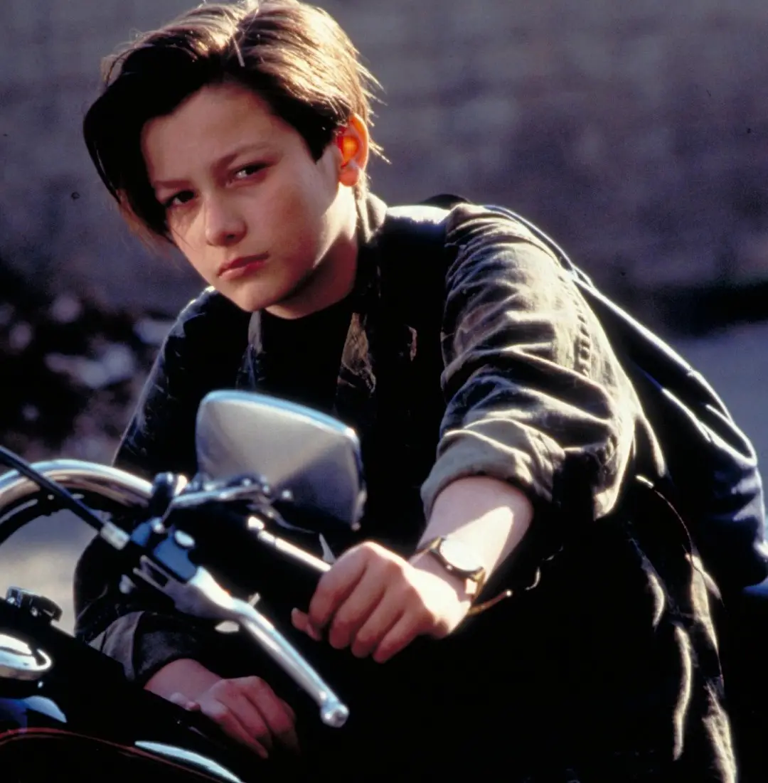 A young boy with dark hair and a serious expression sits on a motorcycle, gripping the handlebars. He wears a dark jacket…