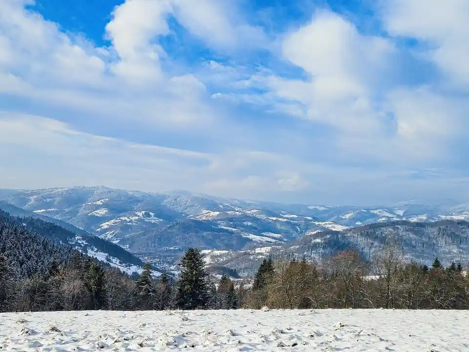 A scenic winter landscape shows snow-covered rolling hills and forests under a bright blue sky with fluffy white clouds. The…