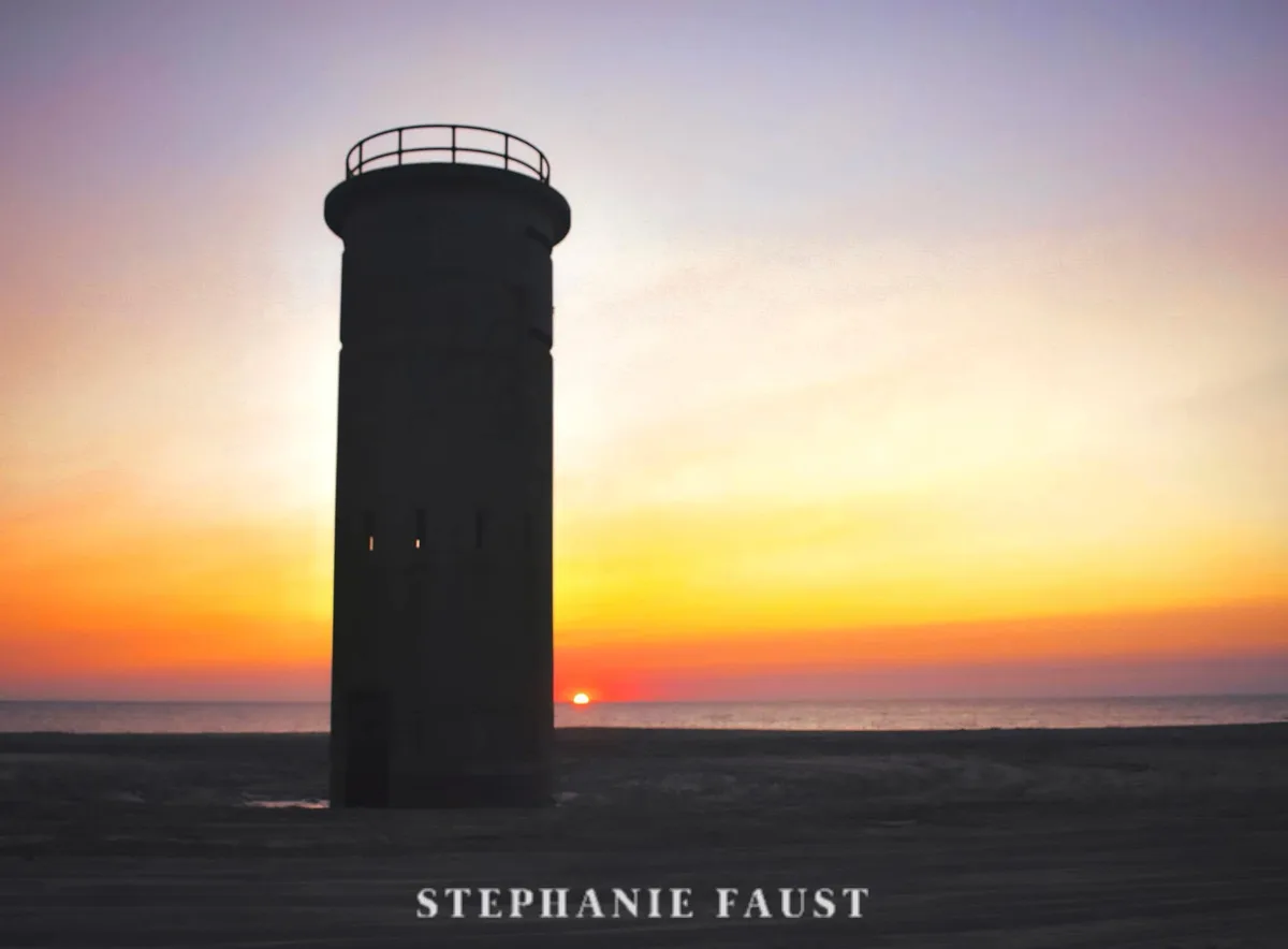 A silhouette of a lighthouse stands on a beach at sunset, with the sun low on the horizon casting orange and purple hues…