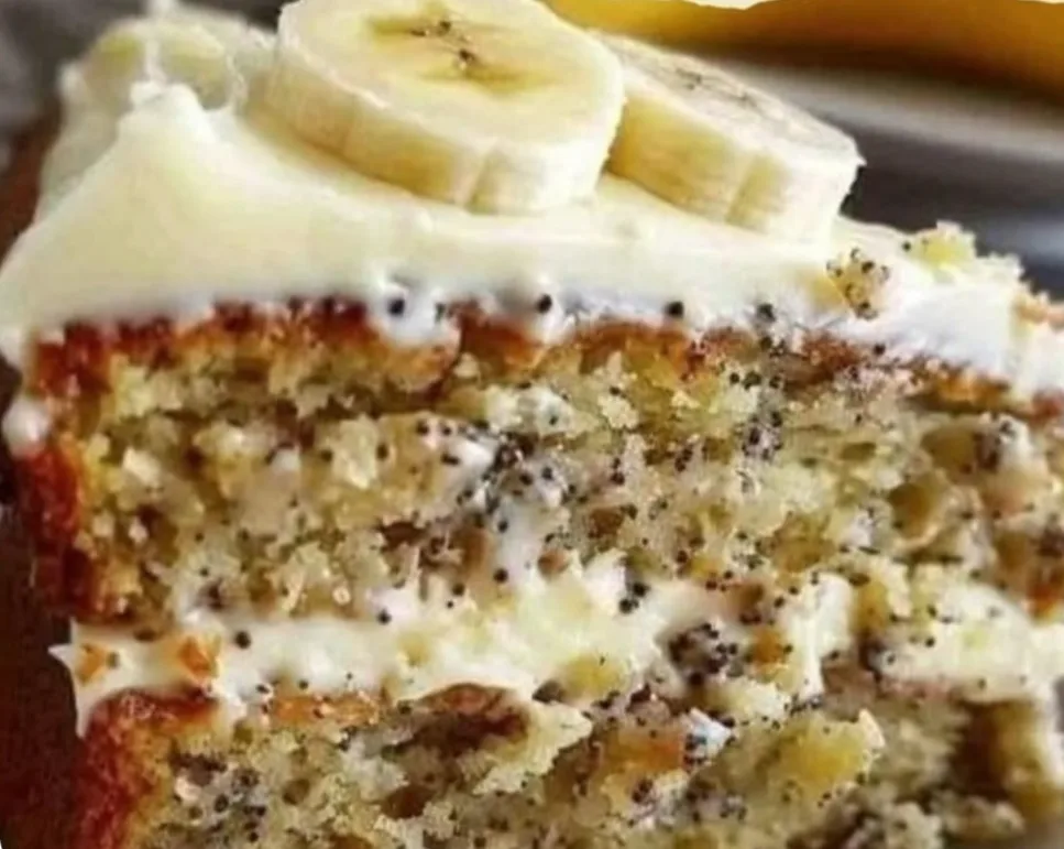 A close-up photo of a slice of banana cake with white frosting and sliced bananas on top, showing the moist cake layers and…