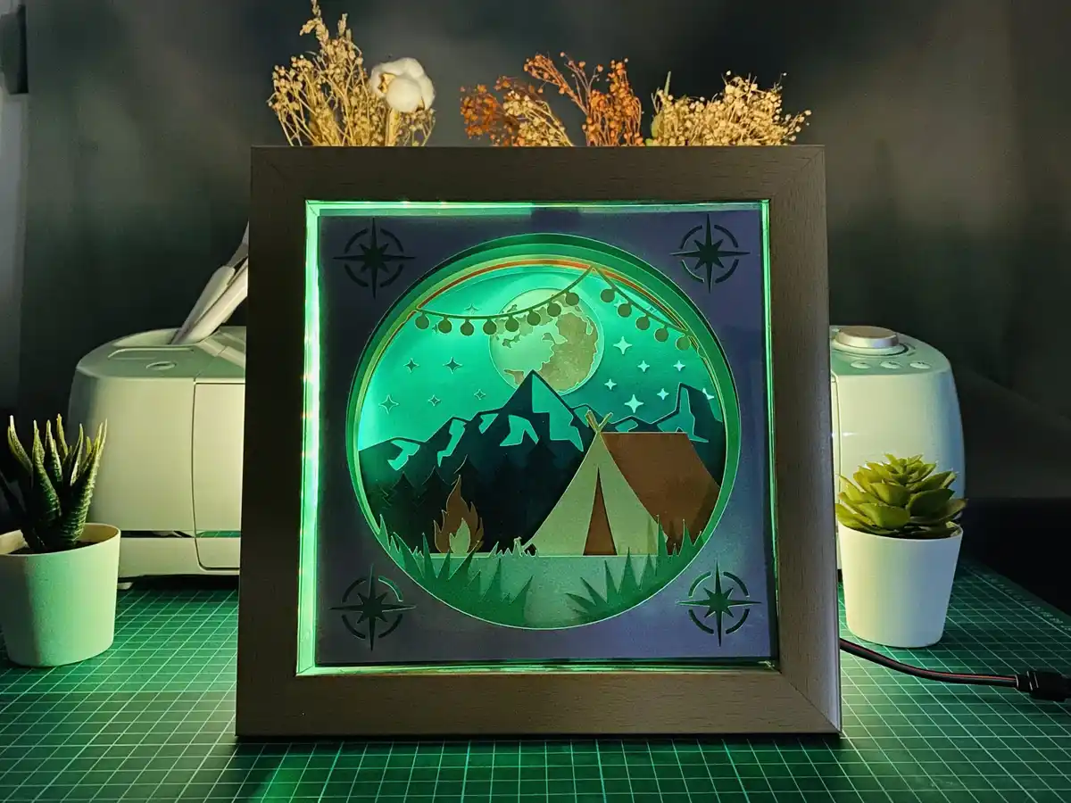 A framed, illuminated 3D paper-cut artwork depicts a camping scene with mountains, a teepee, a campfire, and a glowing moon…