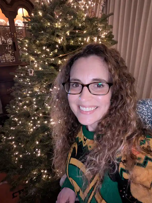 A woman with curly hair and glasses smiles at the camera while wearing a festive green, yellow, and black patterned sweater…