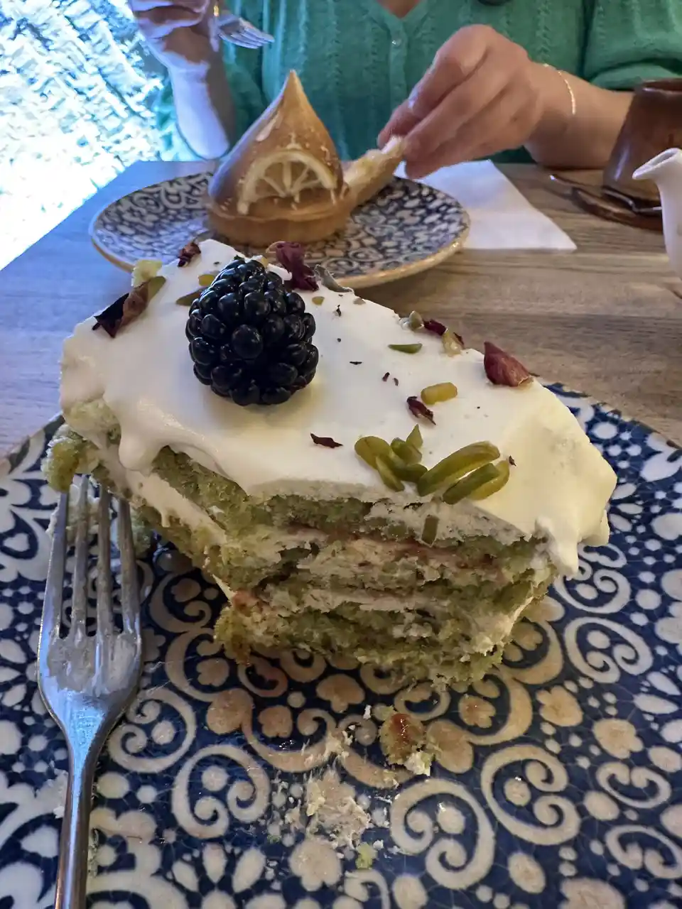 A slice of layered green cake with white frosting, topped with a blackberry and pistachios, sits on a blue and white…
