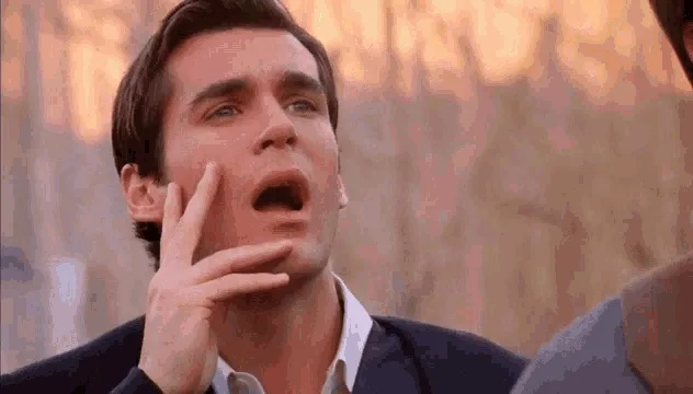 A man with dark hair and a dark jacket looks upward with his mouth open in shock or disbelief, his hand near his face. The…