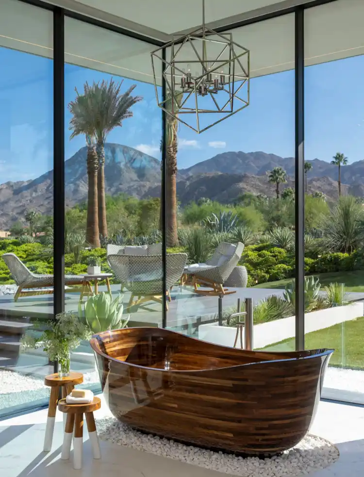 A luxurious wooden freestanding bathtub sits in a modern bathroom with floor-to-ceiling windows overlooking a desert…