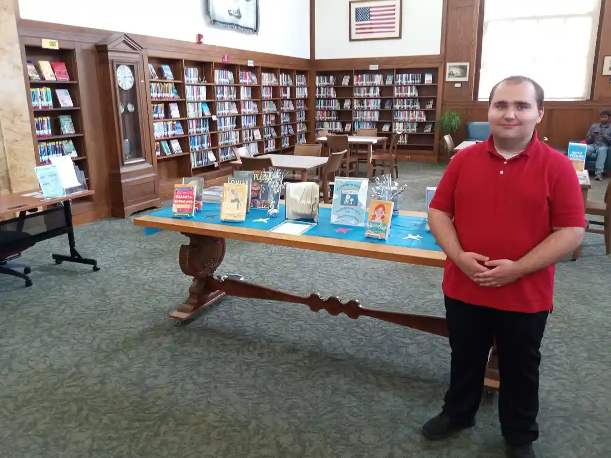 A man in a red polo shirt stands in a library next to a display table with books and decorations. Behind him are wooden…