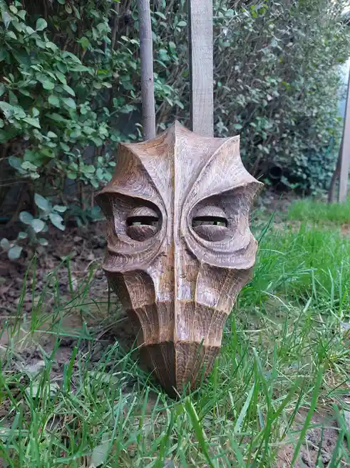 A wooden, intricately carved mask with angular, menacing features and hollow eye sockets is stuck upright in green grass…