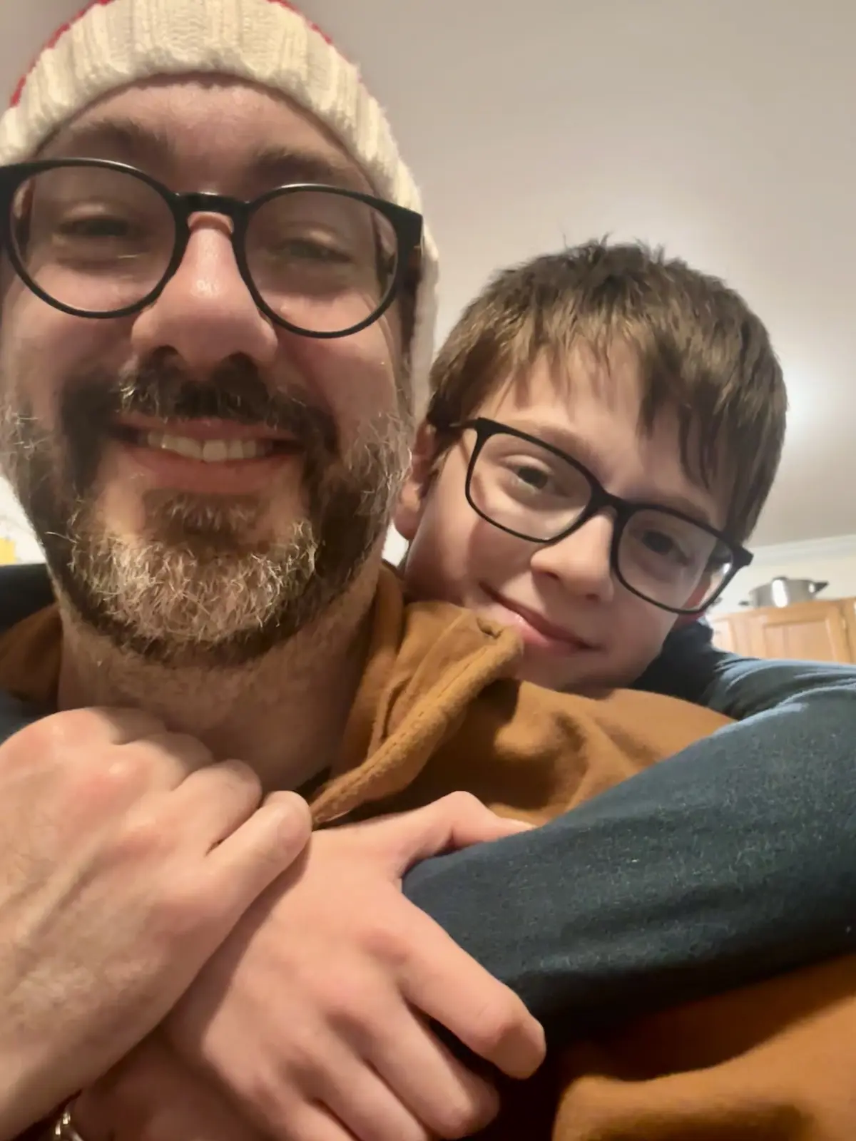 A man with a beard, black-rimmed glasses, and a white knit hat smiles at the camera while being hugged from behind by a…