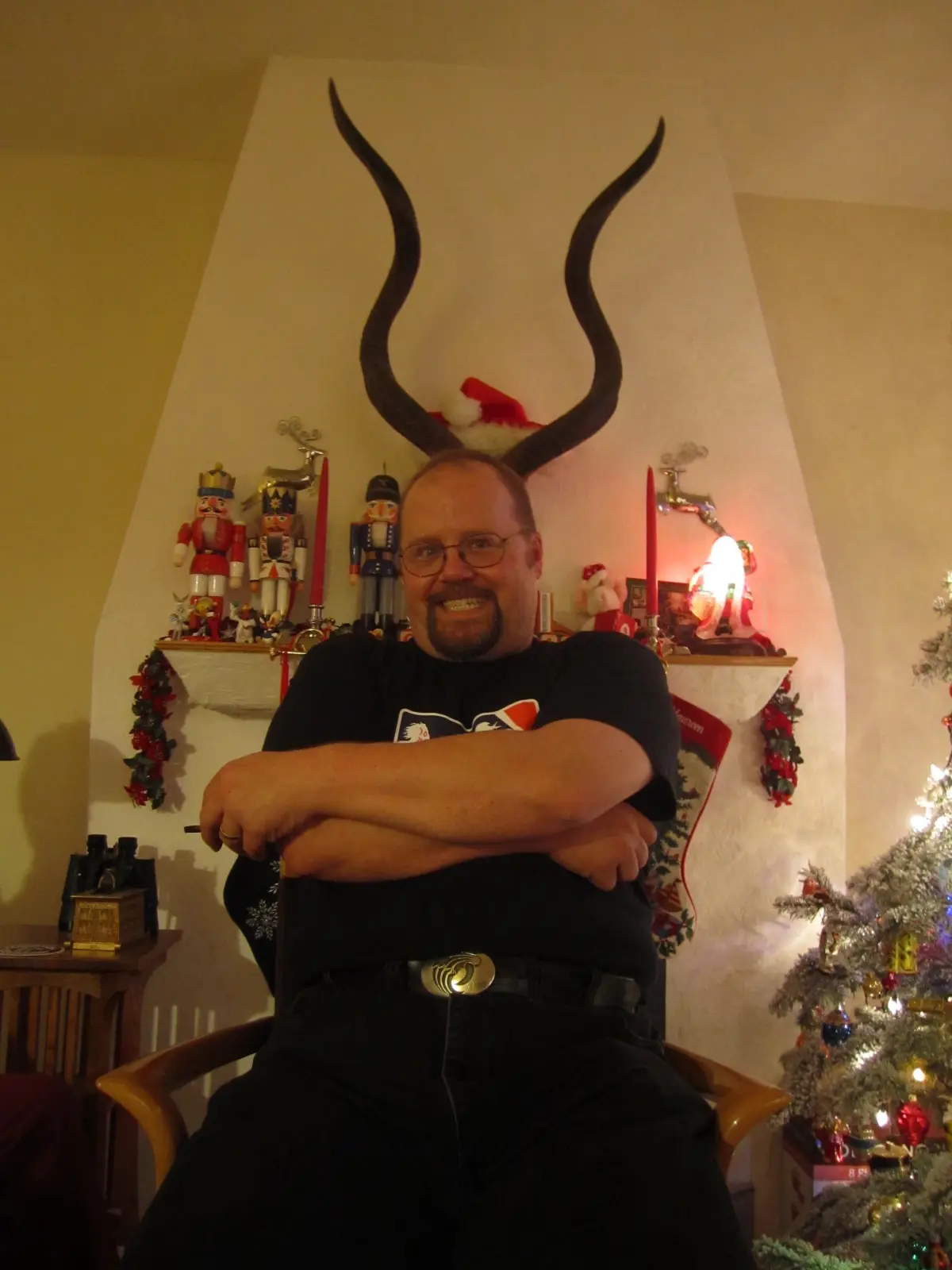 A man with glasses and a beard sits in a chair with arms crossed, wearing a black t-shirt and jeans, with large, curved…