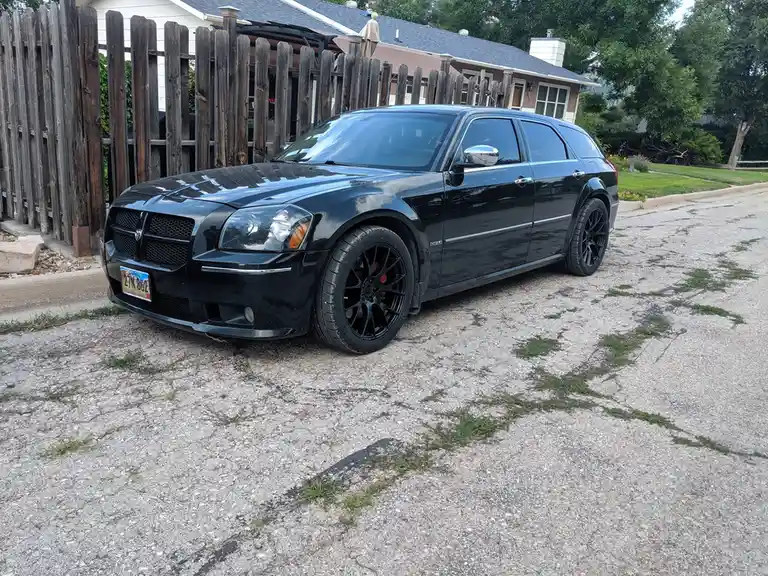A black Dodge Magnum station wagon with aftermarket black wheels and red brake calipers is parked on a cracked, grassy…