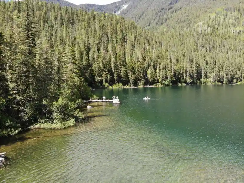 A scenic aerial view of a calm, greenish lake surrounded by dense evergreen forests and mountains. Several people are…