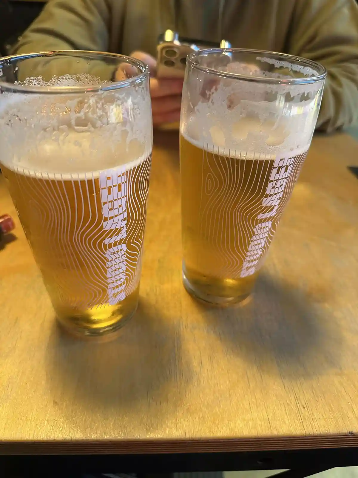 Two tall glasses of beer with white topographic-style patterns sit on a wooden table, partially frothy at the top. In the…