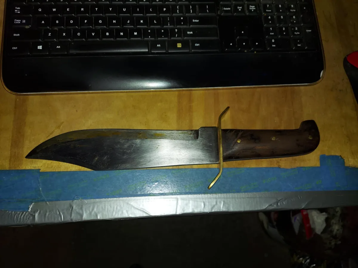A large, worn knife with a dark wooden handle and a metallic blade rests horizontally on a wooden surface, partially atop…
