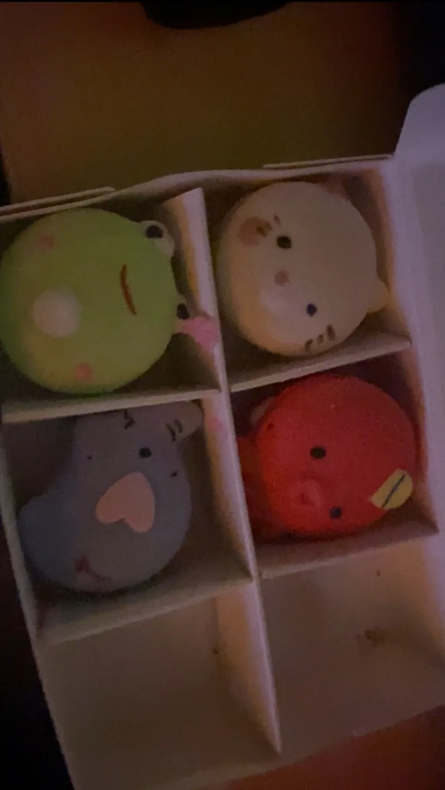 A dimly lit photo shows four small, colorful, round plush toys arranged in a white cardboard box divided into four…