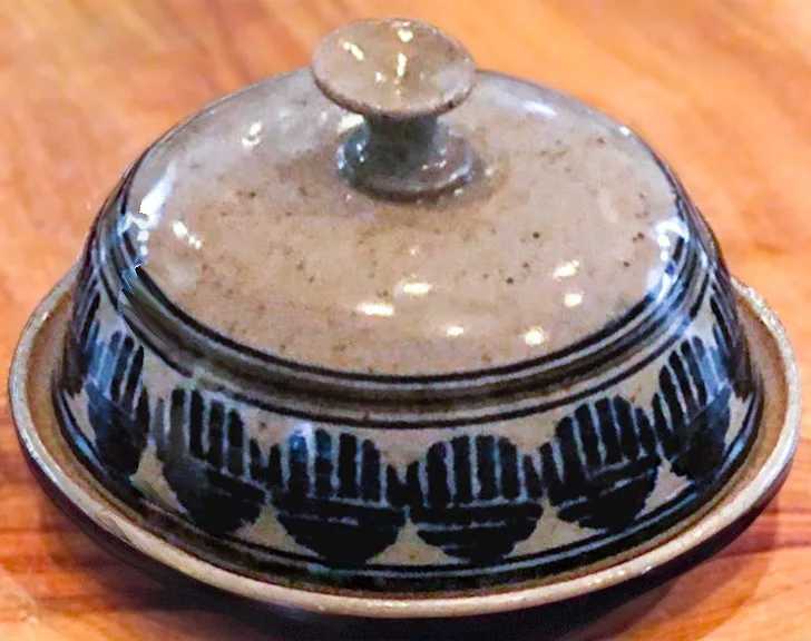 A ceramic butter dish with a lid, featuring a black pattern of stylized hands around its base, sits on a wooden surface. The…