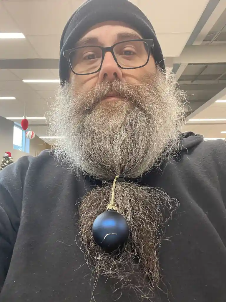 A man with a long, gray beard is taking a selfie indoors, wearing a black beanie and glasses. A black Christmas ornament…
