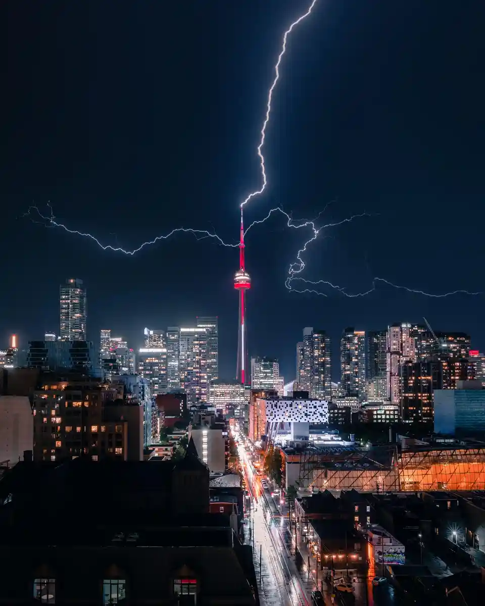 A dramatic night photograph of the Toronto skyline, featuring the iconic CN Tower illuminated in red, struck by a massive…