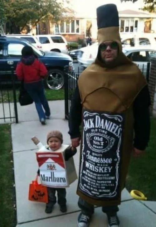 A man dressed in a full-body Jack Daniel's whiskey bottle costume stands holding hands with a small child dressed as a…