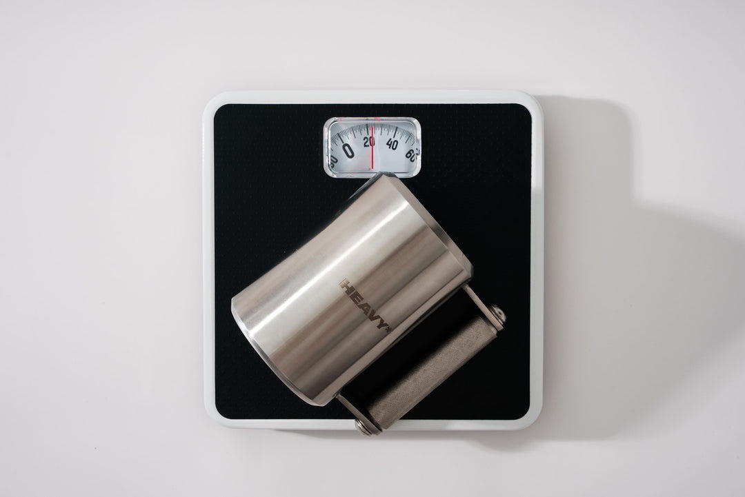 A stainless steel tumbler with the word 'HEAVY' engraved on it is placed on a black analog bathroom scale. The scale's…
