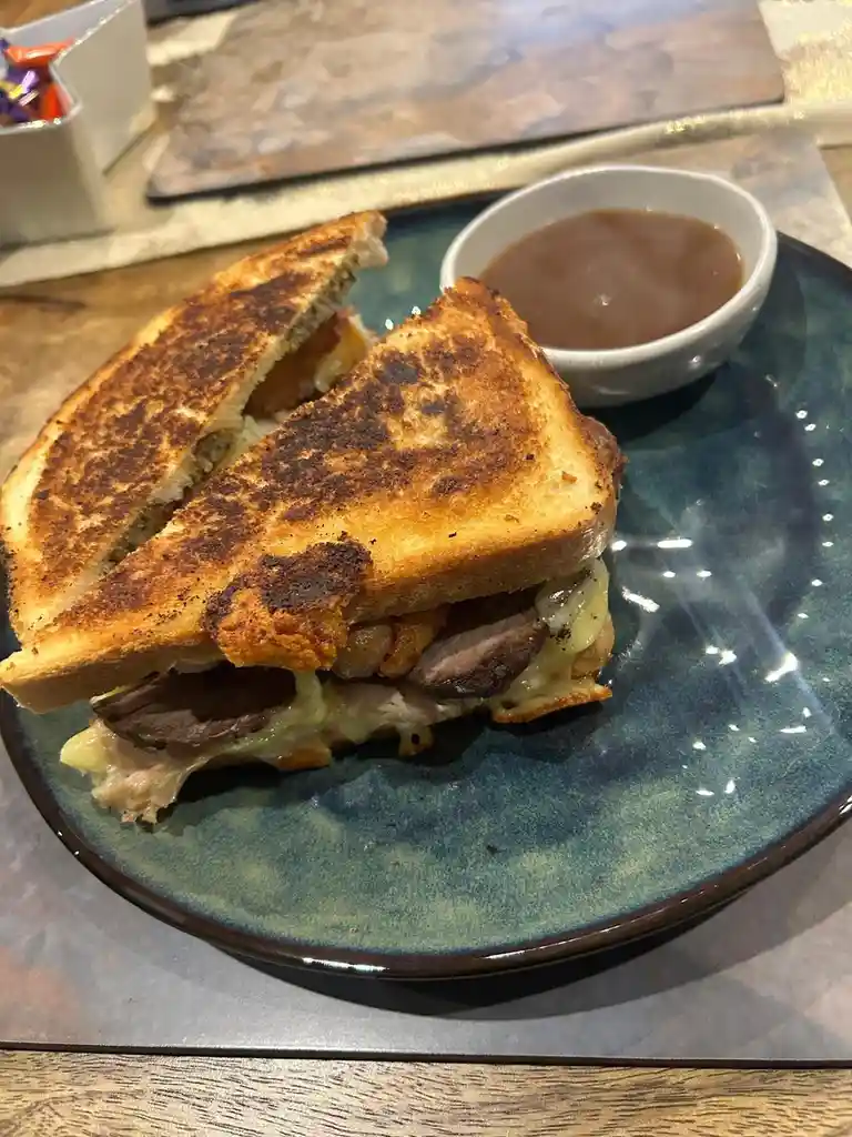 A close-up photo shows a grilled sandwich cut in half, revealing melted cheese and meat inside, served on a blue plate with…