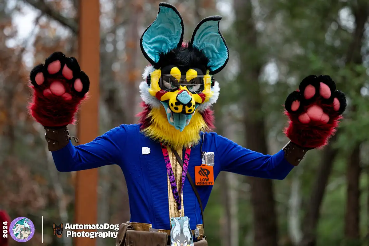 A person in a colorful fursuit with large blue ears, yellow fur, and red paw gloves stands with arms outstretched in a…