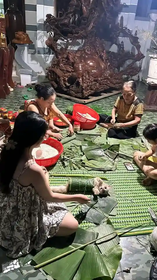 Several people are sitting on a green mat on the floor, preparing food wrapped in large banana leaves. A small, fluffy cat…