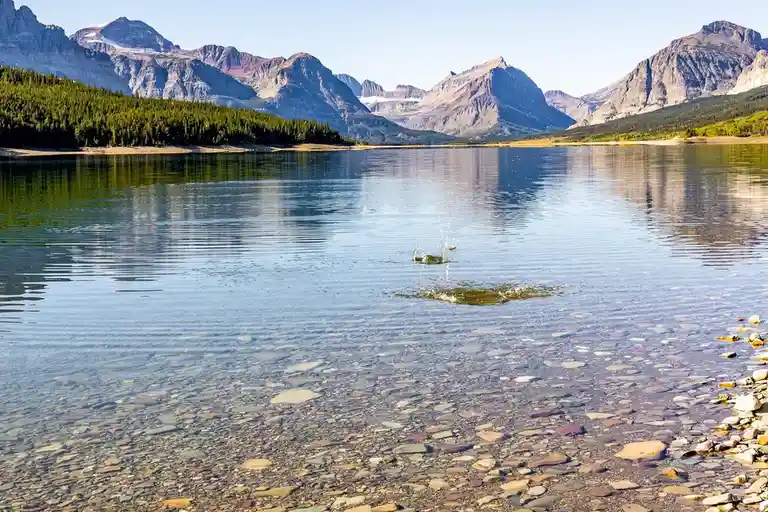 A serene mountain lake with clear water revealing colorful pebbles on the bottom, surrounded by forested hills and…