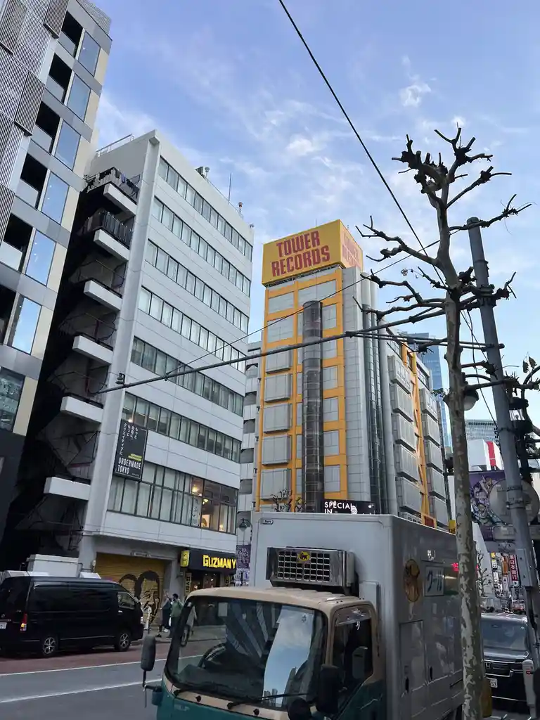 A street view in Tokyo shows a tall building with a large yellow sign reading 'TOWER RECORDS' against a blue sky. A leafless…