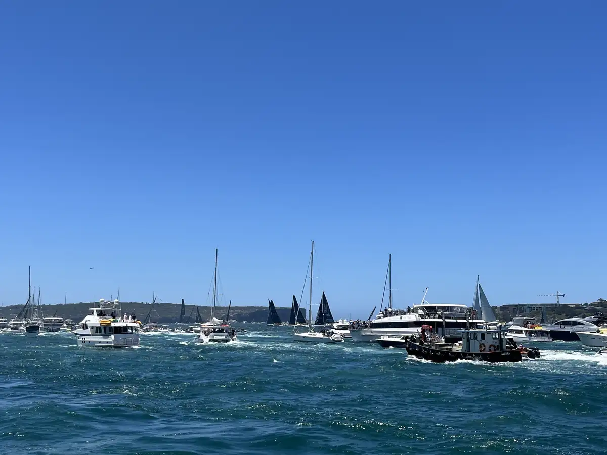 A wide shot of a busy harbor filled with numerous boats, including sailboats with dark sails and motor yachts, all moving…
