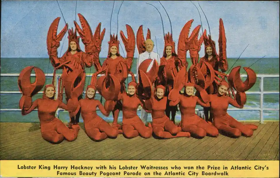 A vintage postcard image shows a group of women in full red lobster costumes posing on a wooden pier with the ocean behind…