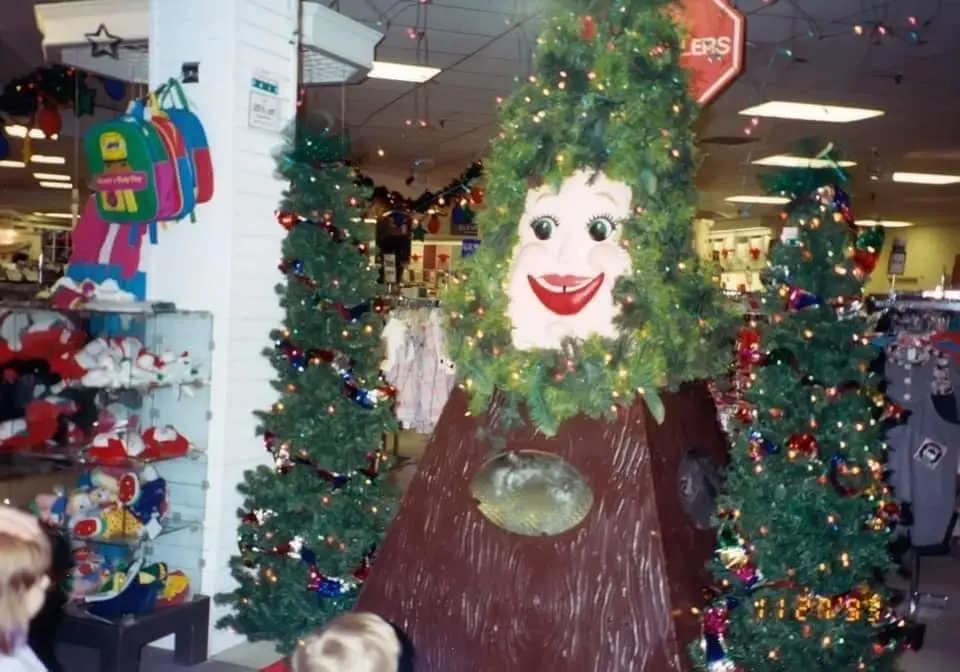 A large, artificial Christmas tree display in a department store, featuring a cartoonish human face with big eyes and a wide…