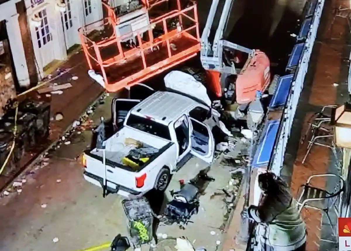 A security camera captures a white pickup truck severely damaged after crashing into a large orange construction lift. The…