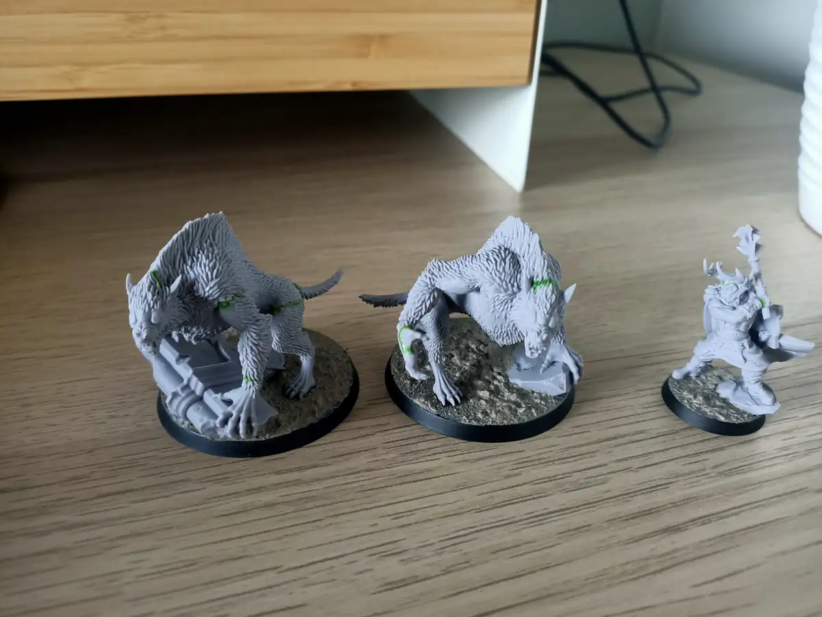 Three unpainted fantasy miniatures sit on a wooden surface: two wolf-like creatures with green paint highlights and one…