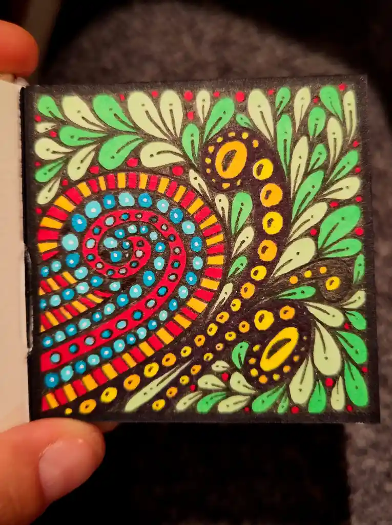 A hand holds a small, colorful, hand-drawn artwork on dark paper featuring a swirling spiral pattern with red, blue, yellow…