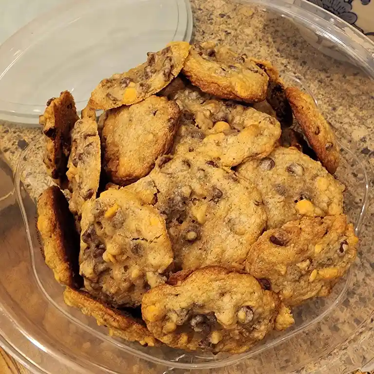 A clear plastic bowl filled with numerous golden-brown chocolate chip cookies, some broken or unevenly shaped, sits on a…