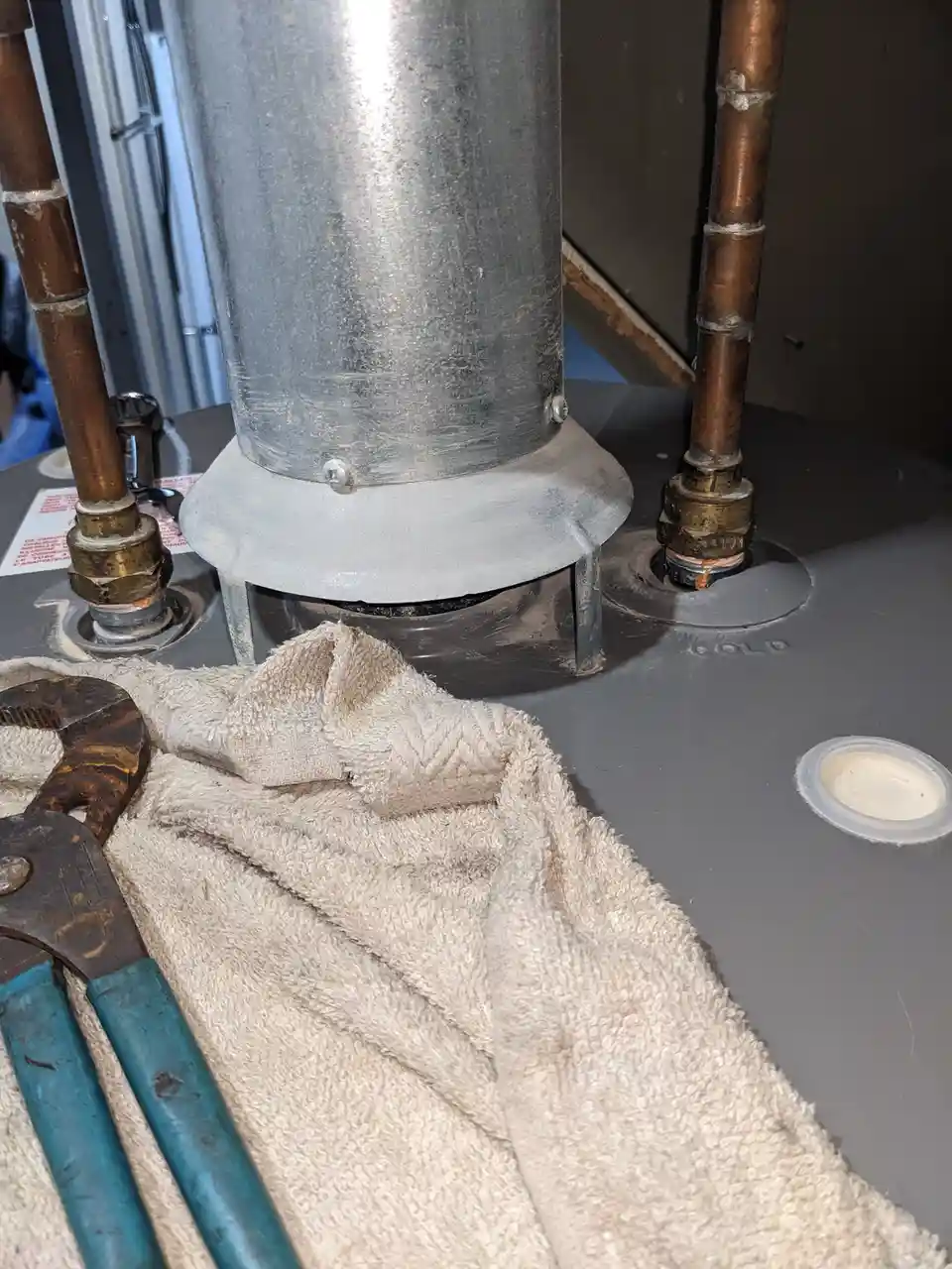 A close-up photo shows a water heater with copper pipes and a metal vent pipe. A rusty wrench and a beige towel lie on the…