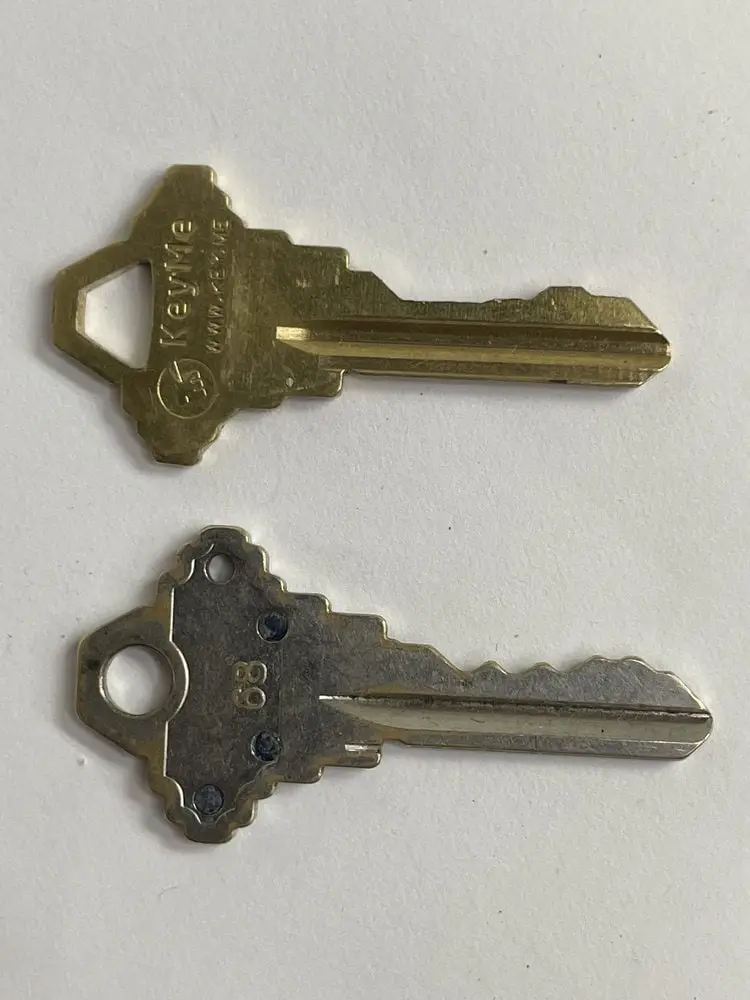 Two keys lie on a white surface; the top one is shiny brass with 'KeyMe' branding, the bottom one is duller metal with '68'…