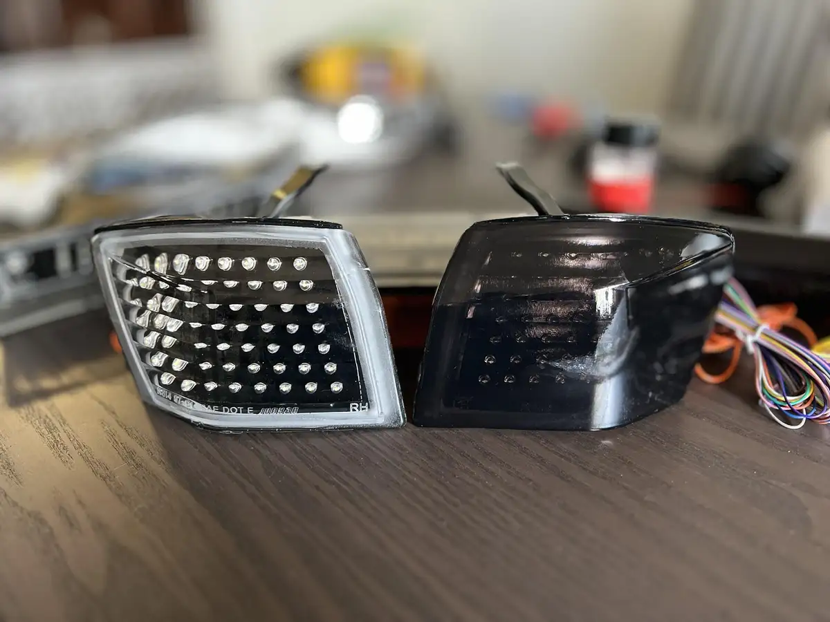 Two car LED headlights sit side-by-side on a wooden table; the left one is clear with visible bulbs, the right one is black…