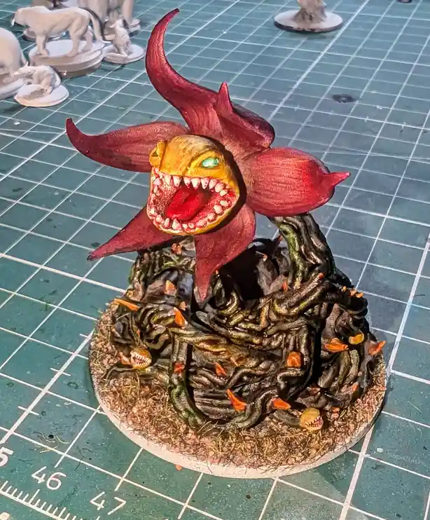 A painted miniature of a monstrous, red, flower-like creature with a gaping mouth full of teeth and a glowing green eye sits…
