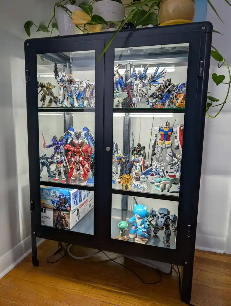 A black glass-front display cabinet filled with numerous Gundam model kits and figures, arranged on multiple shelves. The…